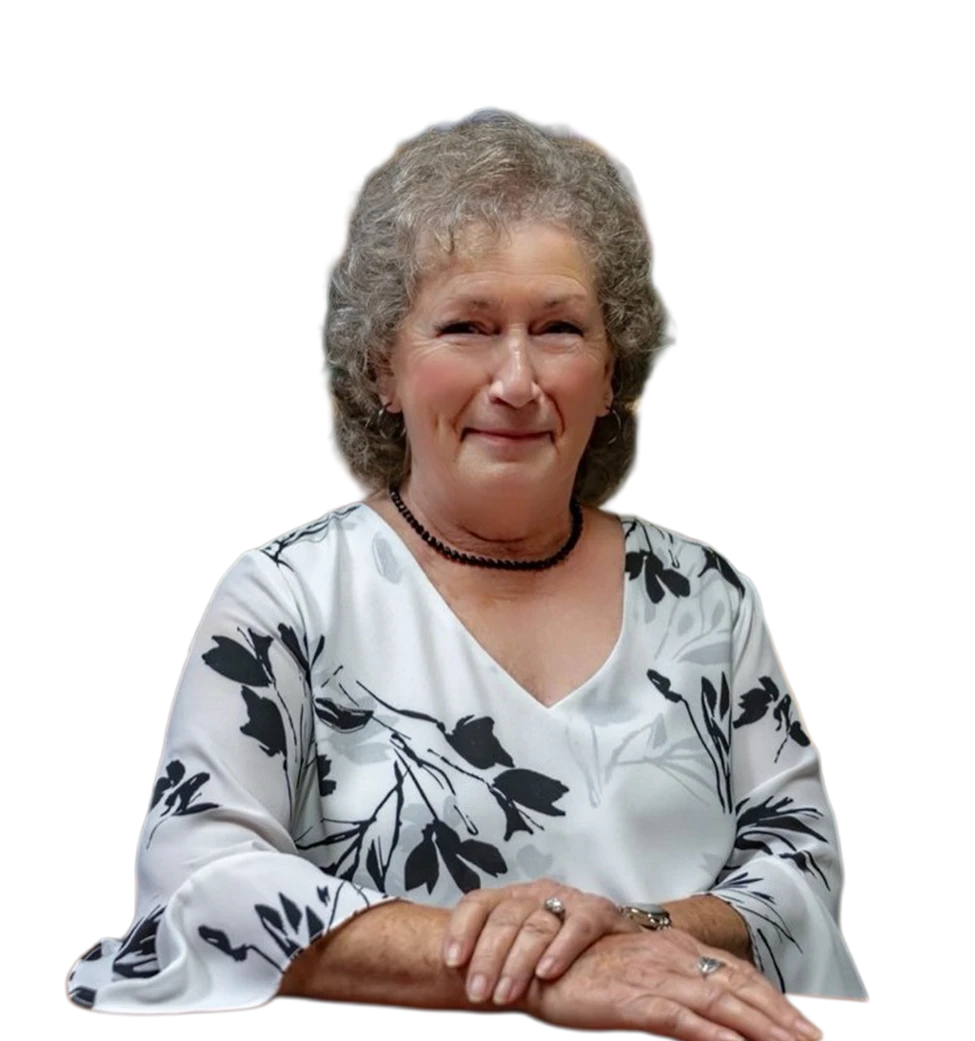 Brenda, smiling with gray curly hair, wears a white floral blouse and black beaded necklace, hands clasped.